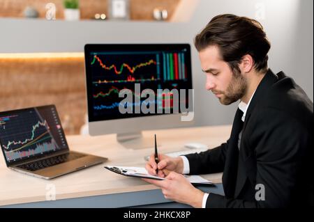 Stock market concept. Focused smart successful caucasian businessman, trader investor, broker, sits at a desk in a modern office, in formal suit, analyze trading charts, predicts profit Stock Photo