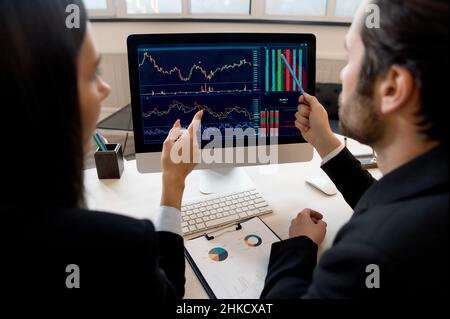 Business colleagues, co-founders the company, managers, stock market traders, sitting at desktop in front of computer screen, analyze the dynamics of charts, discuss strategy, predict, draw up report Stock Photo