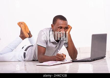 a handsome student in a t-shirt lying on the floor isolated on a white background working using a laptop and taking notes while smiling. Stock Photo