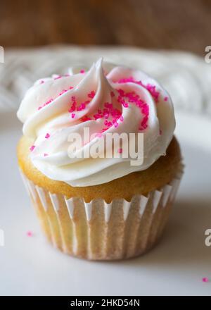 A close up view of a mini cupcake decorated with pink sugar sprinkles.  Confetti cupcake batter with white frosting on a white plate.  Wooden table bl Stock Photo