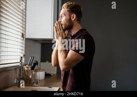Attractive young man looking in the window while standing at the kitchen sink Stock Photo