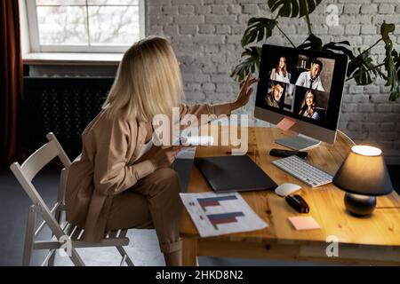 Blond-hair female freelancer using computer, having online meeting with coworkers. Young business woman sitting at the desk in home office, discussing Stock Photo