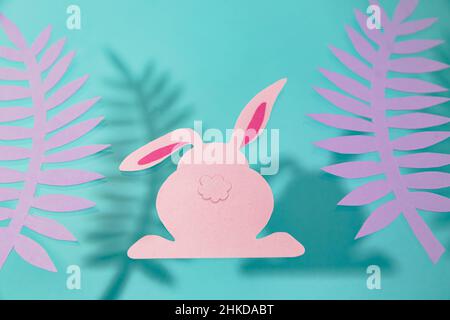 Pink rabbit and palm leaves on a blue background. Paper cut. Easter card. Stock Photo