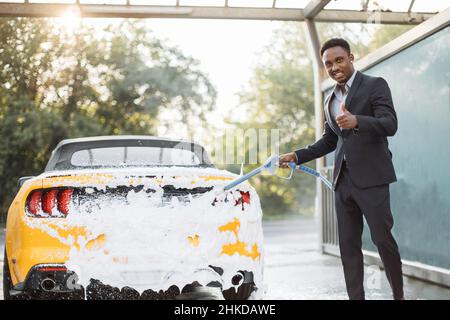 Washing of yellow modern car on open air self car wash service with foam and high powered hose. Young smiling African Confident male entrepreneur cleaning his car with a jet sprayer and soap outdoors.