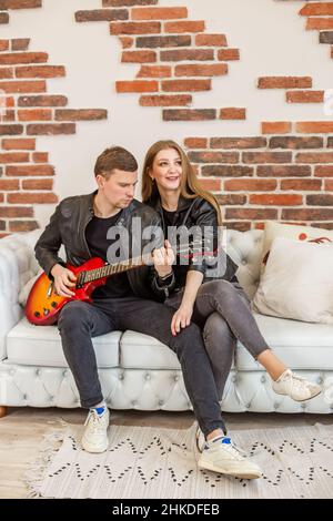 Young couple in love is sitting on sofa. Guy plays rock music on electric guitar. Girlfriend listens to song. Spend time together. Stock Photo