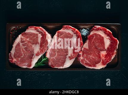 Three fresh raw rib eye steaks on a wooden board on a black background with salt, pepper and rosemary in a rustic style Stock Photo