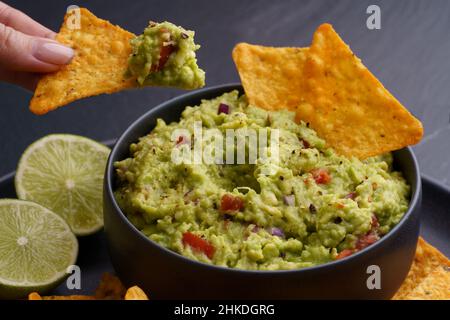 Closeup of woman hand with tortilla chips or nachos with fresh tasty guacamole dip in black plate on black background Stock Photo