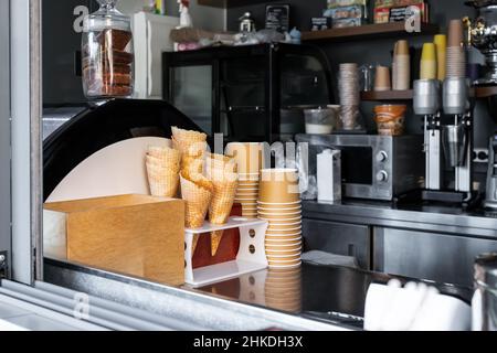 Many empty baked waffle cones and paper cups for ice cream stacked at display counter window of street food takeaway cafe window. Sweet desserts and Stock Photo