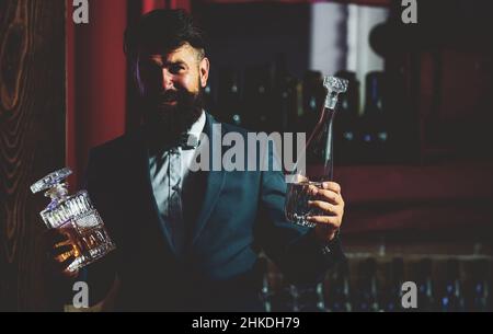 Handsome bearded barman with long beard and mustache with serious face made alcoholic cocktail in vintage suit on bar background. Stock Photo