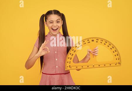 winking teen girl hold school math tool protractor ruler on yellow background show ok, education Stock Photo