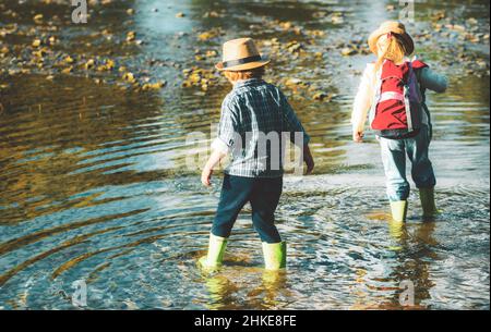 Two kids hiking with backpacks on river. Cute blond girl and boy playing in the creek. Children walking in forest stream and exploring nature. Summer Stock Photo