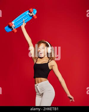 Carefree African American female hipster standing with skateboard in raised hand and listening to music in headphones while looking at camera on red background Stock Photo