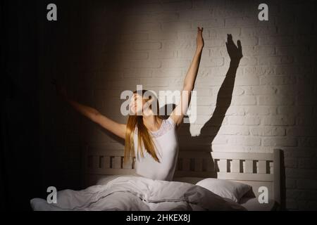 Young woman woke up early in the morning, stretching arms in bed, sunlight breaks through the gap in the curtains Stock Photo