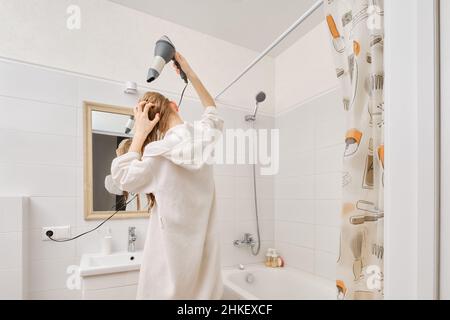 Back view of young woman wearing white bathrobe using hair dryer in bathroom Stock Photo