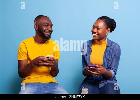 Boyfriend and girlfriend holding mobile phones and smiling at each other, using smartphones with touch screen to browse internet network. Modern couple laughing and using social media. Stock Photo