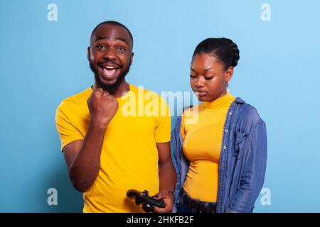 Man beating woman at video games with controller on console over blue background. Boyfriend celebrating win and girlfriend feeling disappointed about losing online gameplay in studio. Stock Photo