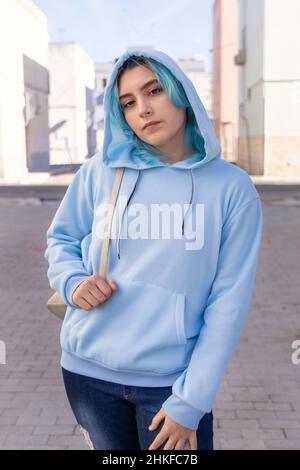 Teenager wearing light blue oversize hoodie and jeans looking into the camera. Blue haired teen girl stays outdoor on city street. Clothing mockup Stock Photo