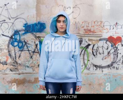 Teenager wearing light blue oversize hoodie and jeans looking into the camera. Blue haired teen girl stays outdoors against graffiti wall. Clothing mo Stock Photo