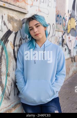 Teenager wearing light blue oversize hoodie and jeans looking into the camera. Blue haired teen girl stays outdoors against graffiti wall. Clothing mo Stock Photo