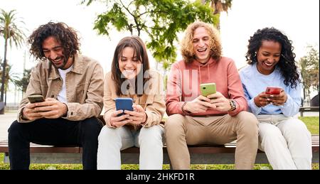 Smiling mixed race group of friends sitting together on bench using mobile phone in the park - friends having fun sharing in social media Stock Photo
