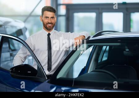 Handsome bearded man in formal outfit visiting auto salon for choosing new vehicle. Happy male customer standing near open door of luxury car and smiling on camera.  Stock Photo