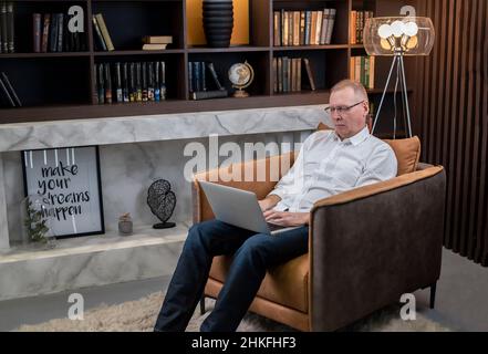 Smart mature businessman in glasses sitting in armchair at home with modern library. Man typing on laptop keyboard. Online work and education concept. High quality photo Stock Photo