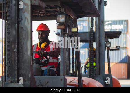 Male worker in uniform and helmet driving and operating on diesel container forklift truck at commercial dock site. Stock Photo