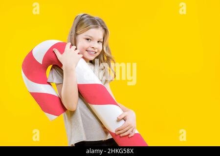 Cute joyful teenage girl holding a huge candy on a yellow background. Happy preschooler with sweets in hands. Lollipop. Christmas sweets. Stock Photo