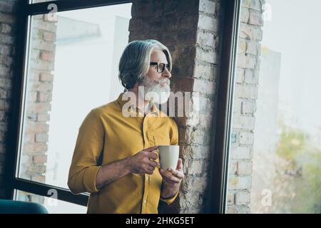 Photo of dreamy senior man wear yellow shirt glasses drinking tasty coffee looking window indoors workplace workstation Stock Photo