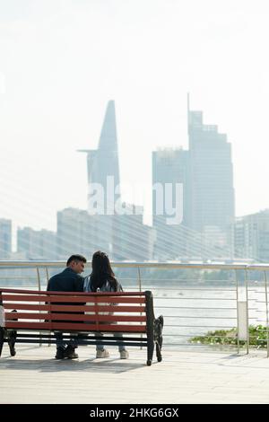 Happy young couple sitting on bench on bridge, talking and enjoying city view Stock Photo
