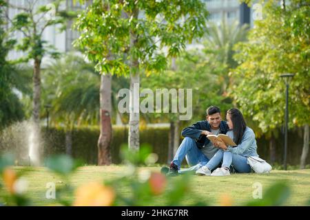 Young university students reading book for class together when sitting on grass on campus Stock Photo