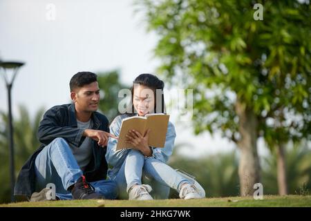 Smiling young couple sitting on grass in park and discussing article in non-fictional book Stock Photo