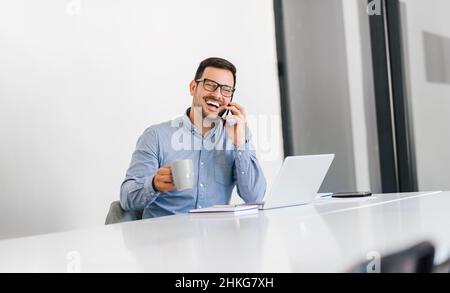 Businessman entrepreneur in casuals drinking coffee talking over mobile phone making smartphone call looking away sitting at desk with laptop in brigh Stock Photo