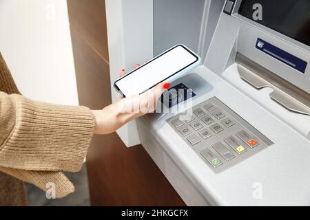 Female hand with mobile phone withdrawing money from atm using NFC contactless wi-fi pay pass system. Wireless authentication and data transmission se Stock Photo