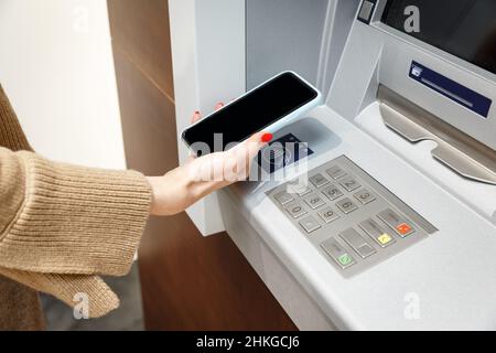 Female hand with mobile phone withdrawing money from atm using NFC contactless wi-fi pay pass system.  Wireless authentication and data transmission s Stock Photo