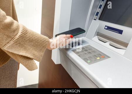 Female hand with mobile phone withdrawing money from atm using NFC contactless wi-fi pay pass system. Wireless authentication and data transmission se Stock Photo