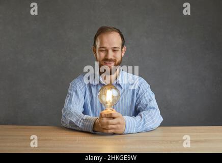 Happy bearded man holding a yellow light bulb sitting at the table on a gray studio background. Stock Photo