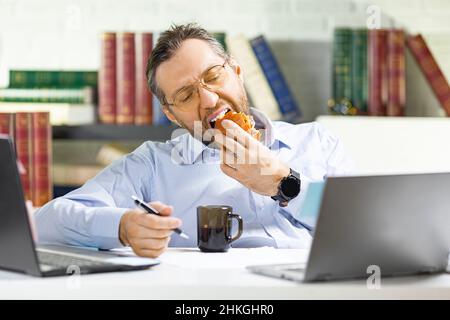 A middle-aged man is lounging in the office - he eats unhealthy food. Stock Photo