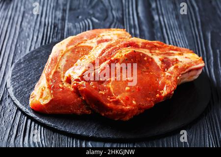 raw bone-in marinated pork chops on a black stone plate on a wood table, horizontal view Stock Photo
