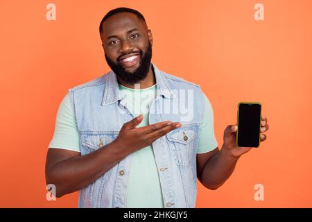 Photo of attractive guy wear denim vest pointing arm modern device empty space isolated orange color background Stock Photo