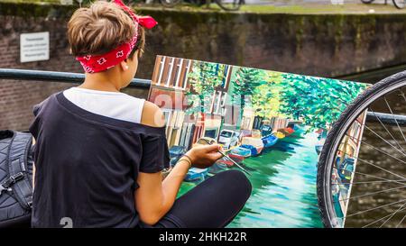 Eine junge Frau im Schneidersitz und rotem Stirnband auf einer Brücke in Amsterdam malt eine typische Häuserfront an einer Gracht