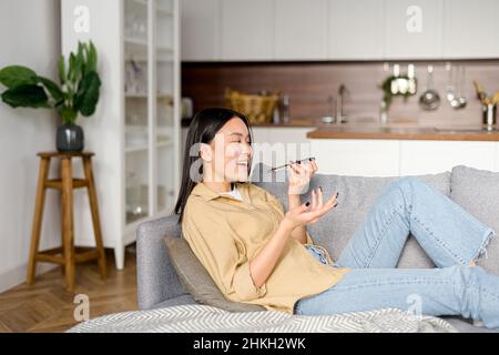 Charming young woman sitting on the sofa at home, recording and sending voice message. Female using voice recognition app on mobile phone, talking in the mic of the smartphone Stock Photo