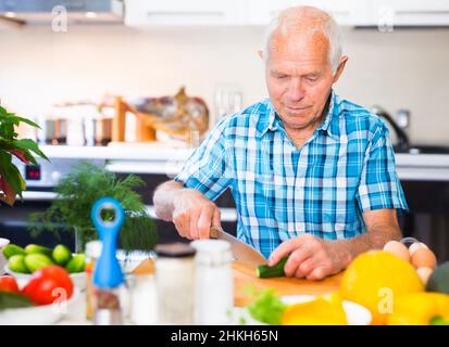 elderly man cuts vegetables for salad at the table in the kitchen Stock Photo