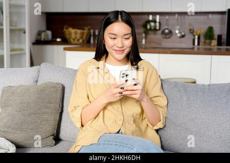Waist up portrait view of the young smiling asian woman sitting at the sofa at home and using smartphone, received unexpected good news. Technologies and people concept Stock Photo