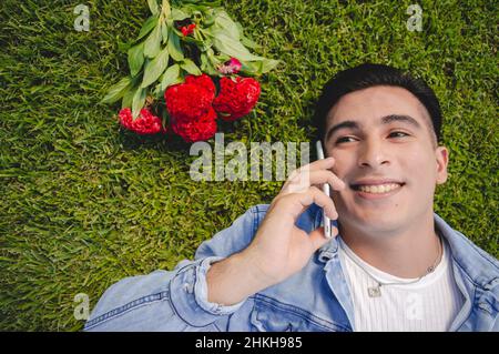 close-up portrait of happy smiling young man communicating by phone with his partner, looking to the side, resting lying on the grass with a bouquet o Stock Photo