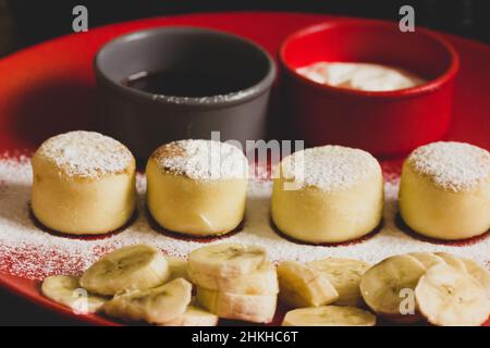 Cottage cheese pancakes or syrniki sprinkled with powdered sugar, served on red plate with sliced bananas, sour cream, jam. Morning breakfast in cafe, Stock Photo