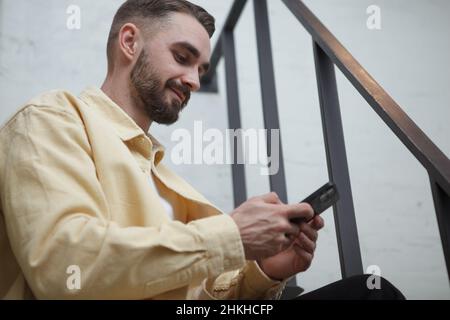 Handsome bearded man smiling, using smart phone app, sitting on the stairs indoors Stock Photo