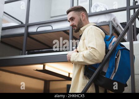 Handsome bearded male traveler walking down the stairs at his hotel room, carrying backpack Stock Photo