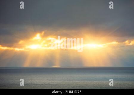 Rays from the sun behind the clouds break through and illuminate the surface of the sea, before sunset Stock Photo