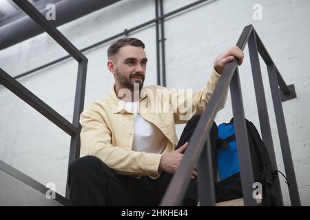 Low angle shot of a handsome bearded male tourist with a backpack sitting on the staircase at hotel room Stock Photo